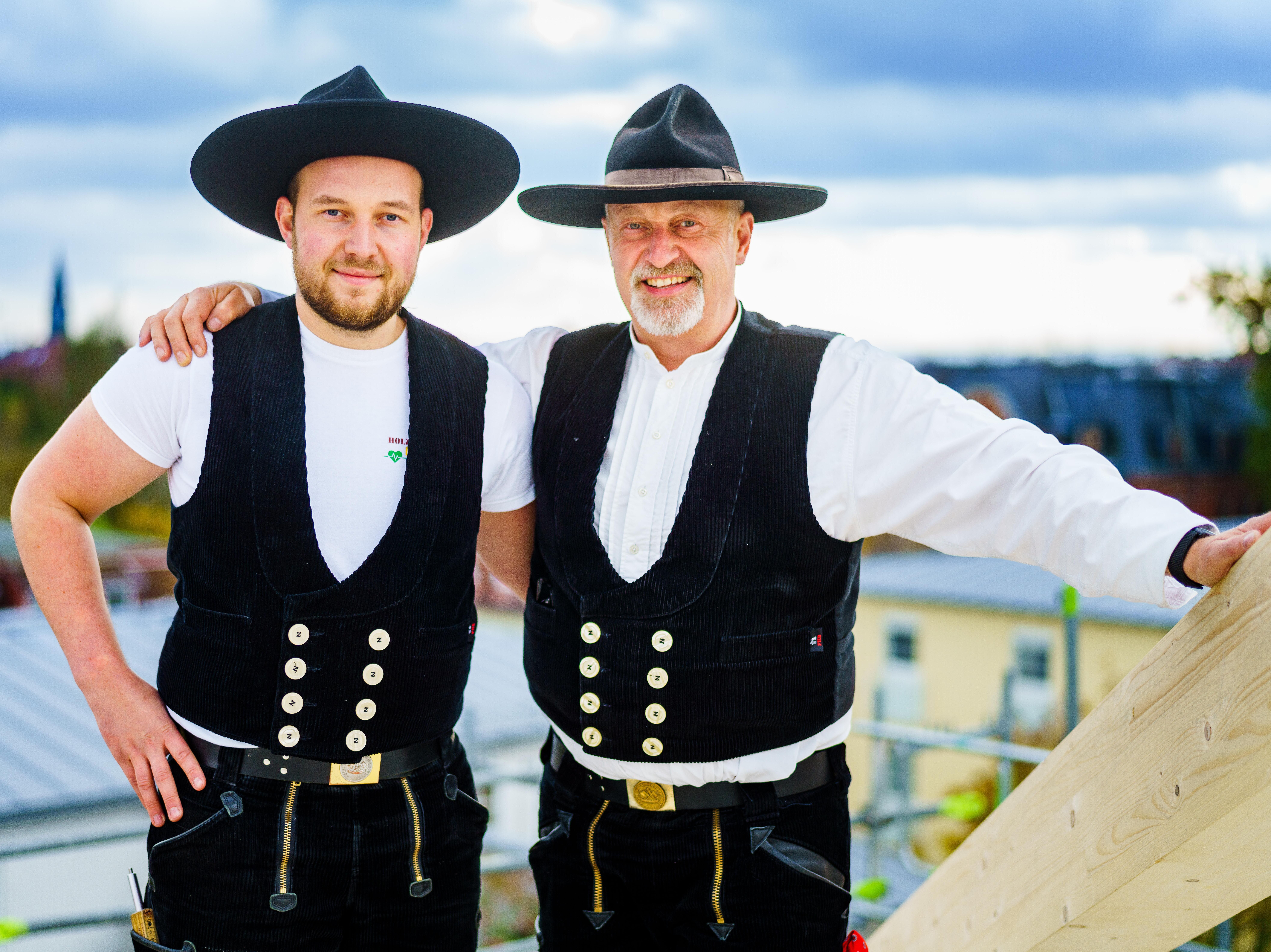 Unternehmensübernahme, Nachfolger Zwei Menschen unterschiedlichen Alters stehen umarmt in Zimmererkleidung auf einem Holzdachstuhl.